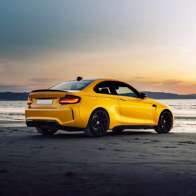 Rear angle of a BMW coupe wrapped in Golden Yellow (RB25-HD) gloss vinyl, perfectly complementing the beachside sunset.
